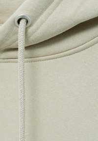 Gros plan du tissu beige d'un sweat à capuche avec cordon tressé inséré dans un œillet métallique près de la couture de la capuche.