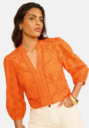 Kobieta o ciemnych włosach nosi pomarańczową bluzkę ażurową z bufiastymi rękawami i kremowe spodnie, uzupełnione złotymi kolczykami kołami i bransoletkami.