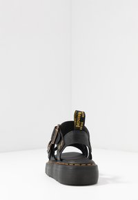 Zwarte sandaal met verstelbare banden, platte zool en gele accenten op de hak. Textuurmateriaal en stiksels zichtbaar op de banden.