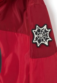 Rode jas met een textuur op de bovenstof, een grote zwart-witte spinnenwebpatch op de mouw en een gladde afwerking overal.