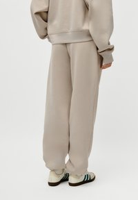 Person som har på seg overdimensjonerte beige joggebukser og matchende genser, parret med hvite joggesko med mørkegrønne striper, stående foran hvit bakgrunn.