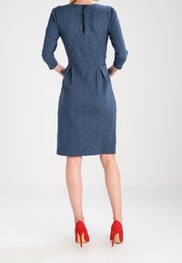 Robe bleu denim avec un motif texturé de feuilles, manches trois-quarts, taille ajustée, côtés plissés, fermeture éclair au dos, et chaussures à talons hauts rouges.