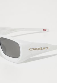 Λευκά γυαλιά ηλίου Oakley με παχιές λεπτομέρειες και σκοτεινούς ορθογώνιους φακούς, που διαθέτουν ένα μεταλλικό λογότυπο Oakley στο βραχίονα του σκελετού.