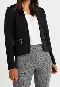 Czarna teksturowana kurtka o kroju cropped z dwoma kieszeniami na zamek, zestawiona z szarymi spodniami w kratę houndstooth i białą bluzką.
