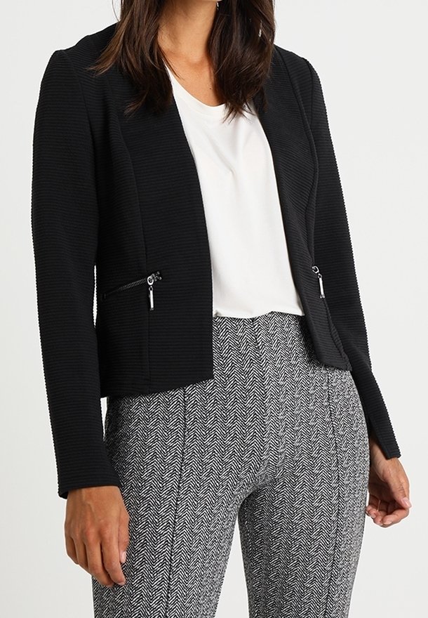 Czarna teksturowana kurtka o kroju cropped z dwoma kieszeniami na zamek, zestawiona z szarymi spodniami w kratę houndstooth i białą bluzką.