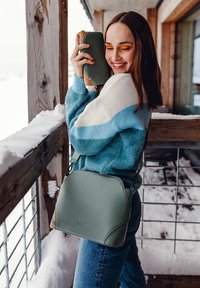 Zelená falešná kožená taška přes rameno se strukturovaným tvarem, kontrastními vzorovanými stranami a nastavitelným popruhem. Měkký modro-bílý svetr.