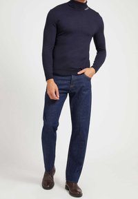 Uomo che indossa un maglione a collo alto blu scuro, jeans blu e stivali in pelle marrone scuro, in piedi con una mano in tasca.