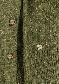 Cardigan verde lavorato a maglia con tessuto testurizzato e riflessi dorati, cinque bottoni marroni e un piccolo etichetta metallica vicino all'orlo.