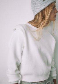 Biely textúrovaný sveter s okrúhlym výstrihom a mierne voľným strihom. Má rebrované manžety a lem. Nosí sa so sivou pletenou čiapkou.