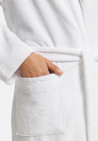 Hvid bambusbadekåbe af frotté med en lomme og bælte. Blød tekstur, lange ærmer og enkelt design. Den ene hånd er placeret i lommen.