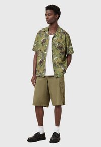 Μπλουζάκι κοντομάνικο, πράσινου χρώματος με μοτίβο από φύλλα φοινικόδεντρου, σε συνδυασμό με ελαιώδεις cargo βερμούδες. Το μοντέλο φορά black παπούτσια και λευκές κάλτσες μέχρι τον αστράγαλο.