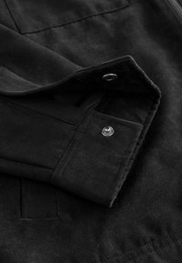 Schwarze Wildlederjacke mit einem Nahaufnahme des Ärmels, der über einen Druckknopfverschluss verfügt und eine weiche Textur sowie saubere Nähdetails zeigt.