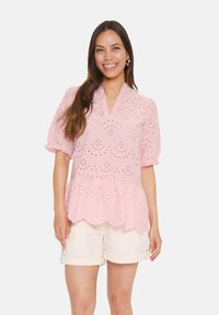 Blusa rosa pallido con maniche corte a sbuffo, dettagli in pizzo e orlo scalloppato. Abbinata a pantaloni corti di colore chiaro.