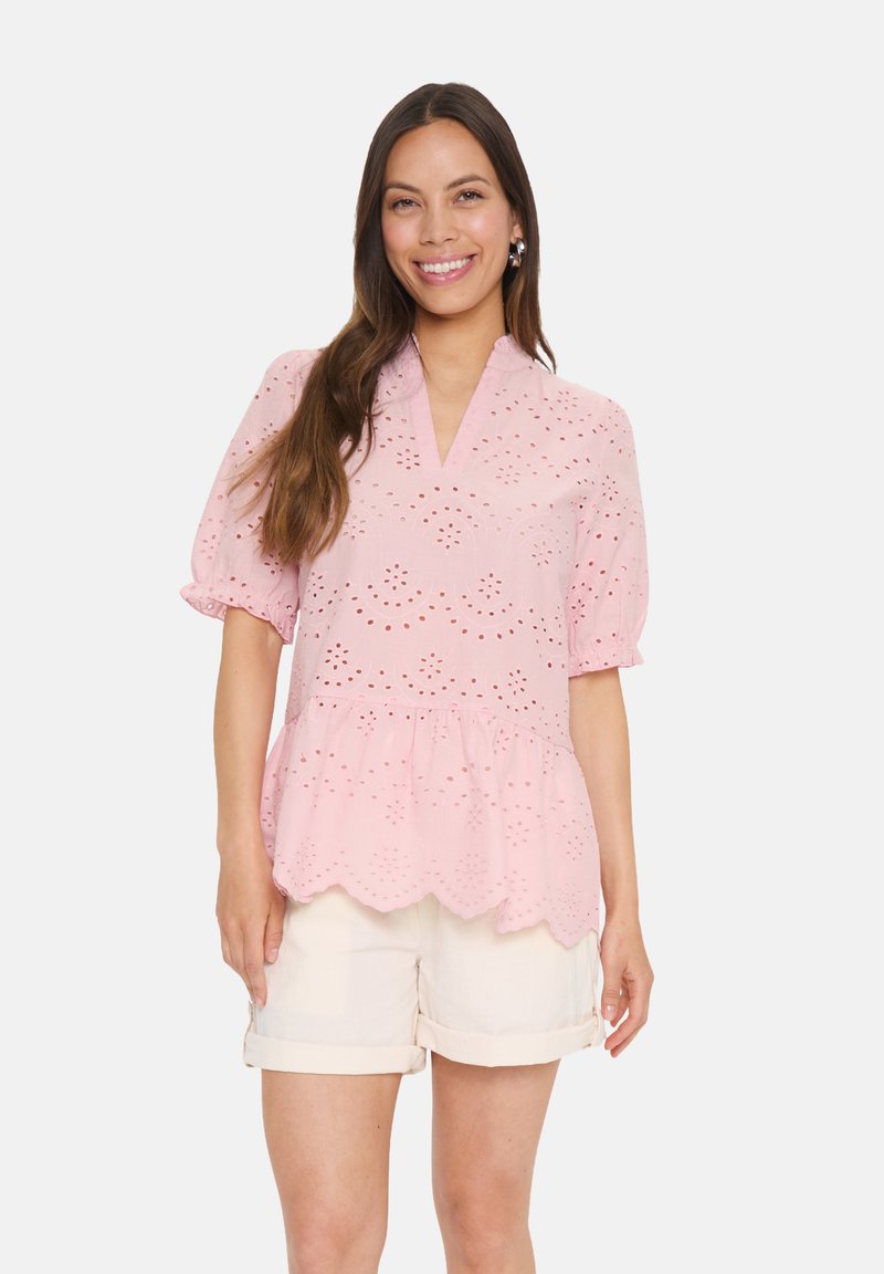 Blusa rosa pallido con maniche corte a sbuffo, dettagli in pizzo e orlo scalloppato. Abbinata a pantaloni corti di colore chiaro.