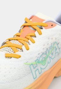 Baskets de course blanches avec une tige en mesh texturé, des accents orange, des lacets jaunes vifs et une semelle rembourrée arborant la marque HOKA.
