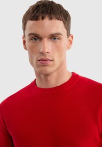 Czerwony sweter z prążkowanej dzianiny z okrągłym dekoltem, charakteryzujący się gładką fakturą i dopasowanym krojem, skupiający się na torsie, na neutralnym tle.