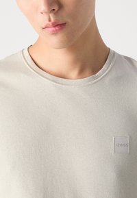 Jasnoszary t-shirt z okrągłym dekoltem, wykonany z gładkiej bawełnianej tkaniny. Posiada małe wytłoczone logo "BOSS" na lewej piersi. Prosty design.