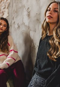 Twee individuen zitten tegen een textuurwand. De een draagt een zwarte trui en een pantalons met een patroon; de ander heeft een crème trui met roze strepen aan.