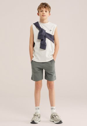 Niño de pie mirando hacia adelante, vistiendo una camiseta sin mangas blanca, pantalones cortos grises, zapatillas deportivas y una sudadera oscura atada sobre el pecho con las manos en los bolsillos.