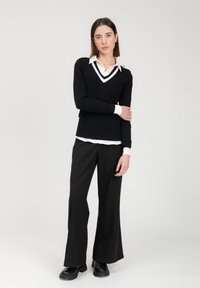 Schwarzer Strickpullover mit weißem V-Ausschnitt und Schulterknöpfen, über einem weißen Hemd mit Kragen getragen. Lockere schwarze Hose und schwarze Stiefel.