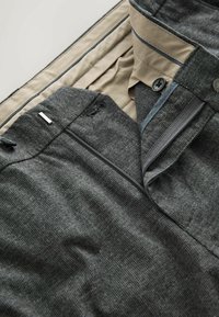 Harmaat kudotut housut, joissa on teksturoitu pinta, beige vuori, vetoketjukiinnitys ja nappi-yksityiskohta. Vyötäröllä on koristeellinen metallikoriste.