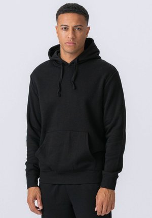 Mężczyzna w gładkiej czarnej bluzie z kapturem z przednią kieszenią i troczkami, stojący na tle jednolitego jasnego tła.