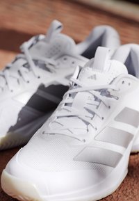 Een paar witte Adidas sportschoenen met grijze strepen en veters, geplaatst op een bruine gestructureerde ondergrond.