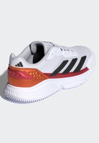 Witte sportschoen met textuurstof, zwarte strepen, een oranje-rode tussenzool en een geribbelde witte zool voor grip.