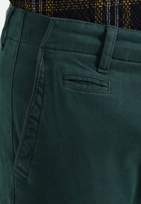 Gröna bomullsbukser med en platt bakficka med ett horisontellt snitt, tillverkade med robust sömnad och en slät yta.