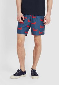 Pantaloni da bagno blu con un motivo a ripetizione di bassotti rossi, cintura elastica e tasche laterali. Abbinati a sneaker blu scuro.