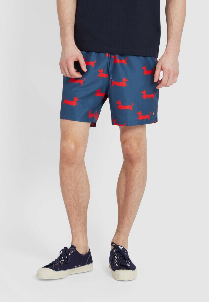 Pantaloni da bagno blu con un motivo a ripetizione di bassotti rossi, cintura elastica e tasche laterali. Abbinati a sneaker blu scuro.