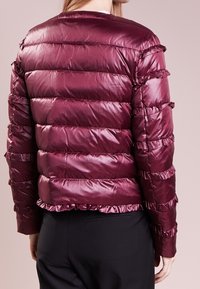 Burgundy pufferjakke med volangdetaljer, horisontal quiltning og cropped design. Glat, skinnende tekstur med elastisk kant i kanten.