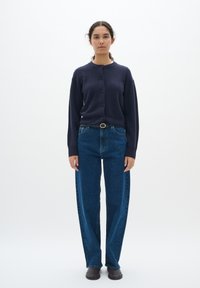 Marinblå kofta med knappar, vida blå jeans och mörka skor. Koftan har rund hals; jeansen har en klassisk denimtextur.