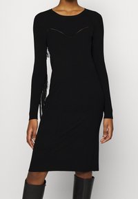 Robe noire en tricot côtelé à manches longues, avec une découpe en V profonde et des accents de franges le long d'un côté. Texture lisse.