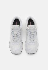 Harmaat mokkanahkakengät, joissa on nauhakiristys. Varustettu pehmustetulla nilkkasaumalla, mustalla sisäpohjalla ja valkoisella välipohjalla, jossa on New Balance -logo.