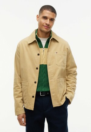 Mand iført beige cordjakke over grøn og beige kvartlyn sweater og marineblå bukser, stående med den ene hånd i lommen mod hvid baggrund.