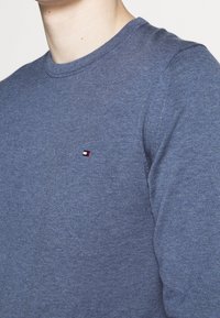 Maglione blu a collo rotondo in maglia con un piccolo logo rosso, bianco e nero sul lato sinistro del petto, con colletto e polsini a costine.