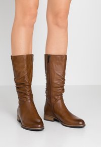  bottes montantes marron en cuir avec un design froissé, fermeture éclair sur le côté, et un talon bas empilé. Texture lisse, accents minimalistes.