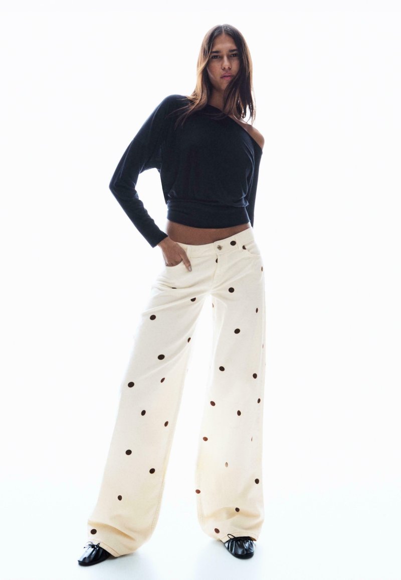 Fekete váll nélküli felső, krémszínű, bőszárú nadrággal, nagyméretű barna pöttyökkel díszítve. A anyag könnyűnek tűnik, sima textúrájú.