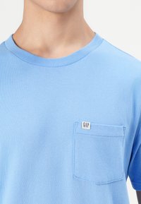 Világoskék, rövid ujjú pamut póló. Kerek nyakkivágással és egy mellzsebből áll, amelyen a "GAP" felirat található.