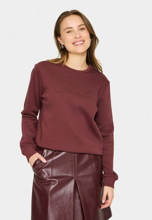 Le sweat-shirt maron présente un col rond, des manches longues et du texte brodé. Il est associé à une jupe en cuir bordeaux brillante avec des plis.