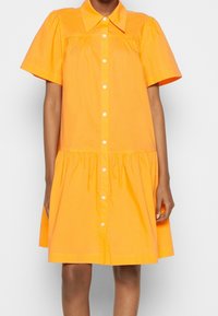 Robe chemise en coton orange avec des manches bouffantes courtes, fermeture à boutons sur le devant et jupe froncée. Elle possède un col pointu et une coupe longueur genou.