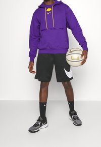 Sweat à capuche Nike violet avec accents jaunes et une poche zippée à l'avant, associé à un short noir avec le logo Nike blanc. Chaussures de sport noires et grises.