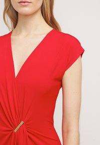 Robe rouge avec un décolleté en V et des manches courtes, présentant un détail torsadé à la taille et une petite accentuation dorée. Texture de tissu lisse.