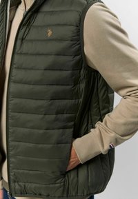 Maslinasto zeleni puffa prsluk s kviltanim dizajnom, koji sadrži zatvarač i grb. Nosiv preko svijetlo bež majice s dugim rukavima.