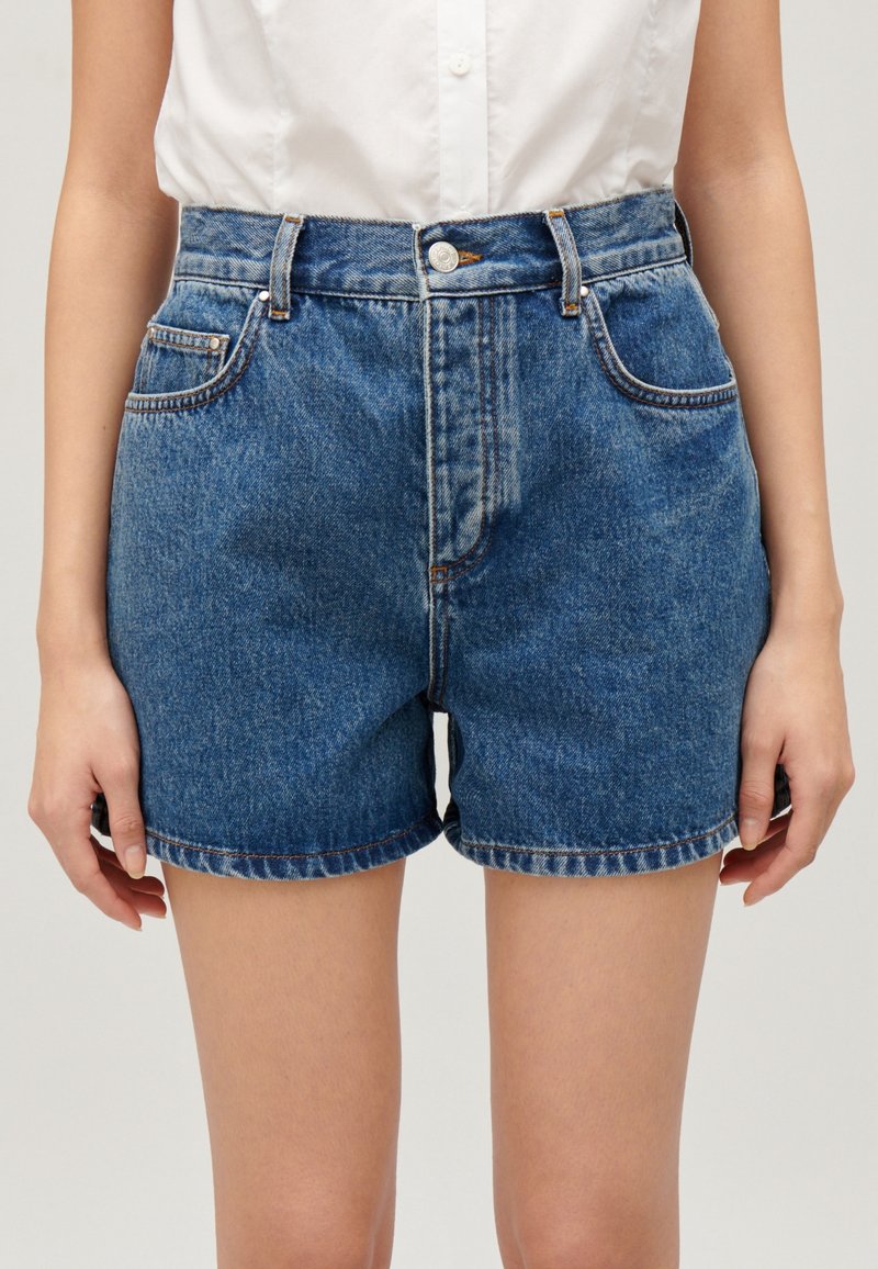 Szorty dżinsowe w średnim niebieskim odcieniu, z wysokim stanem, zapinane na guziki, z pięcioma kieszeniami i podwiniętym brzegiem. Gładka tekstura z minimalnym blaknięciem.