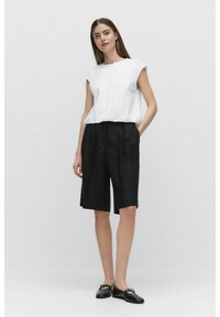Hvid ærmeløs top kombineret med knælange sorte shorts. Stoffet synes let og glat, suppleret af sorte flade sko.