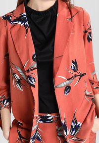 Korálový blazer s květinovým vzorem v modré a bílé barvě, zkombinovaný s černým topem. Má volný střih a texturovanou látku.
