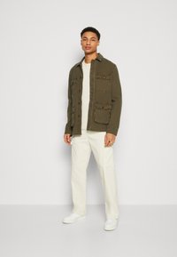 Chaqueta verde oliva con cierre de botones, cuatro bolsillos frontales y un acabado ligeramente texturizado. Combinada con pantalones beige claros y zapatillas blancas.