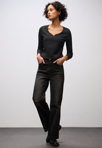 Schwarzes, geripptes Langarmoberteil mit V-Ausschnitt, kombiniert mit hochgeschnittenen, locker sitzenden schwarzen Jeans. Das Model trägt schwarze Knöchelstiefel.